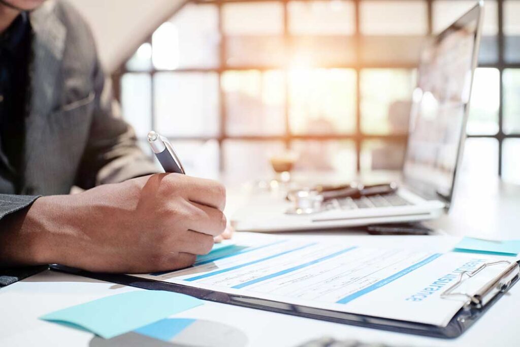 Adult reviewing health insurance documents beside an insurance card and laptop at a kitchen table.